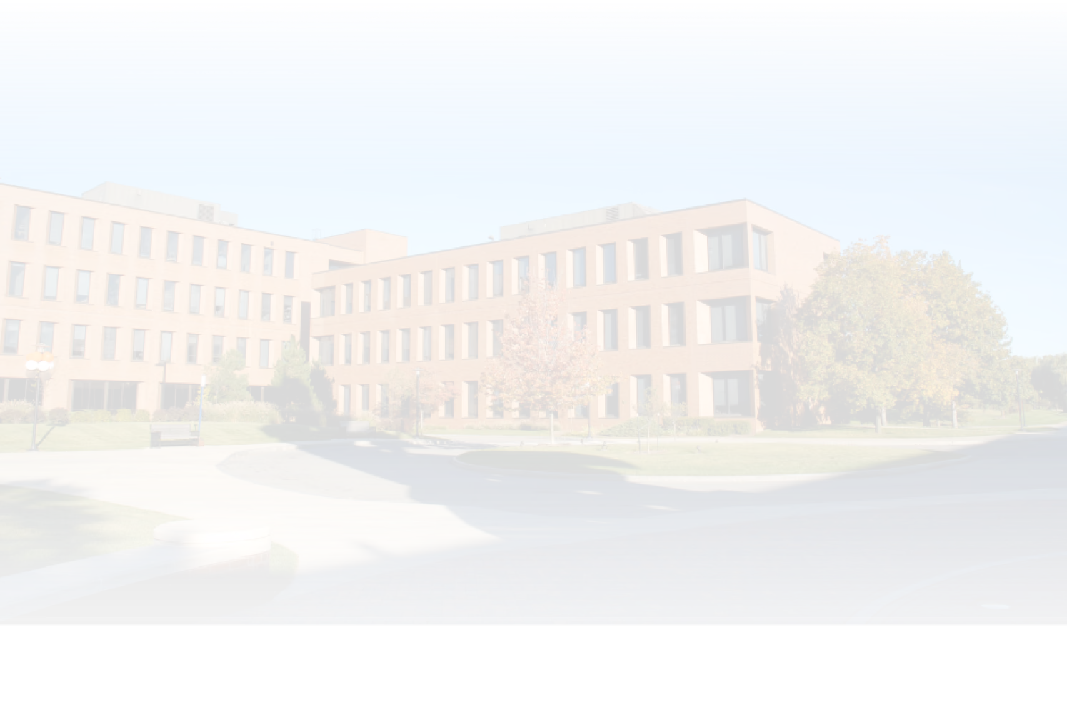 Wide view of a three-story brick college campus building with many windows, surrounded by green lawns and trees under a clear blue sky.