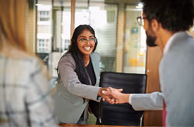 Smiling woman in business attire shaking hands with a man in an office setting.