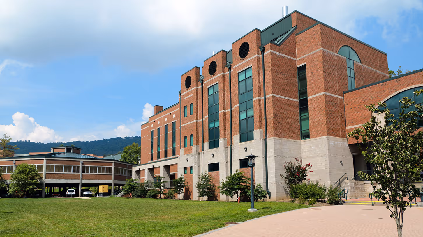 Large brick university building with tall vertical windows and a green lawn in front under a partly cloudy sky.