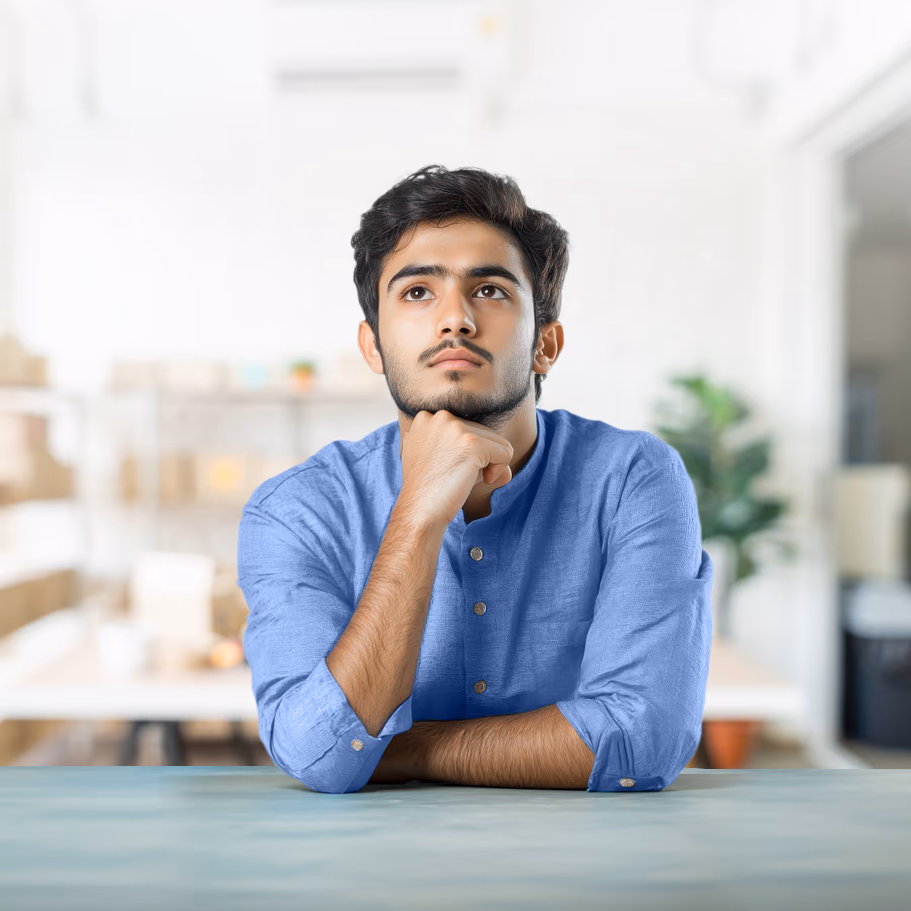 Young man in a blue shirt resting his chin on his hand, looking thoughtfully upward.