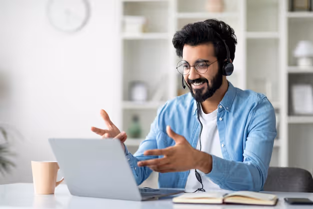 Man with glasses and headset smiling and gesturing while video calling on laptop at desk.