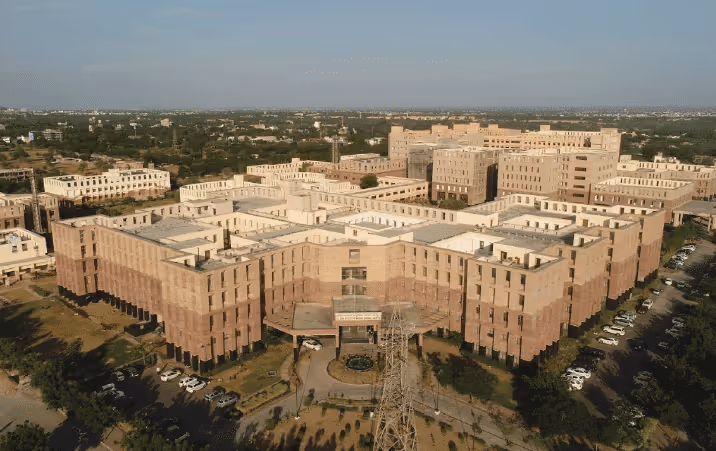 Aerial view of a large, multi-wing institutional building complex with light-colored rooftops and surrounding greenery.