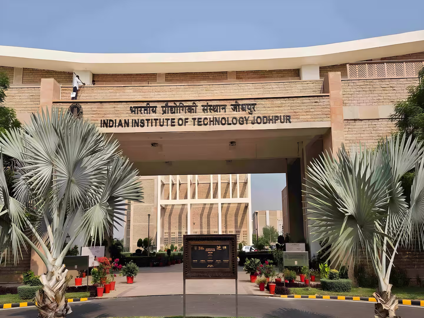Entrance arch of the Indian Institute of Technology Jodhpur with palm trees and potted plants in front.