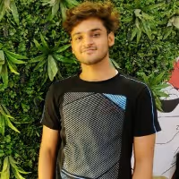 Young man with curly hair wearing a black and gray patterned t-shirt standing in front of green leafy plants.