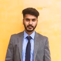 Man with dark hair and beard wearing a gray suit, light blue shirt, and dark tie standing against a yellow background.