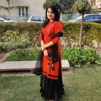 Woman in a red and black traditional outfit standing on grass in a garden with trimmed hedges and parked cars behind her.
