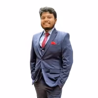 Man wearing a dark blue suit with a maroon tie and pocket square, smiling with hands in pockets.