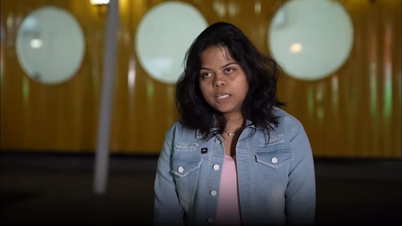 Young woman with shoulder-length dark hair wearing a light blue denim jacket and pink shirt speaking indoors with circular lighted windows behind her.