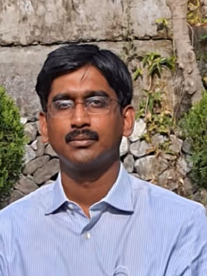 Portrait of a man with glasses, a mustache, and short black hair, wearing a light blue striped shirt, standing outdoors in front of stone walls and greenery.