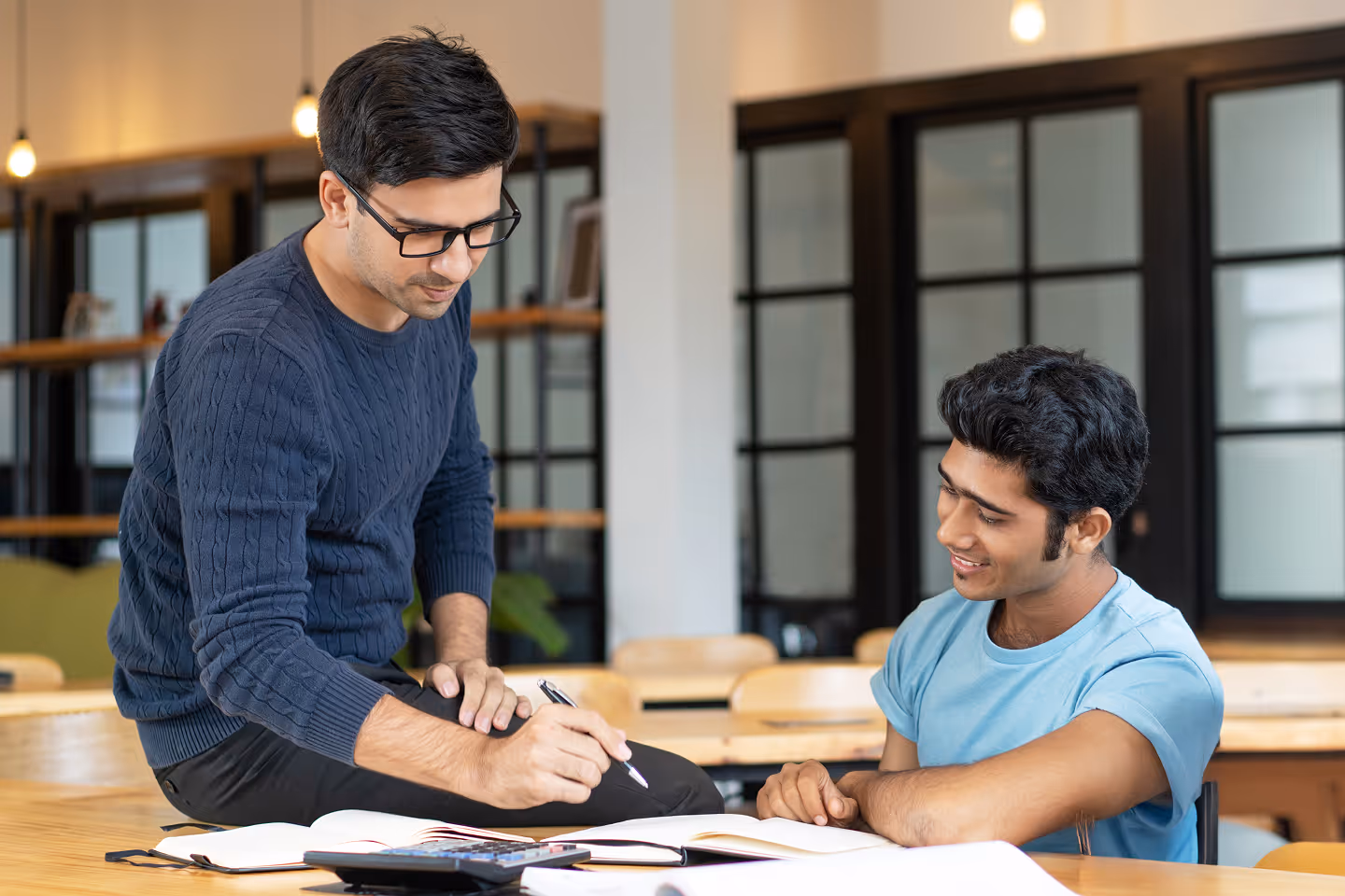 Two young men studying together in a classroom, one sitting and pointing at a book while the other listens and watches.