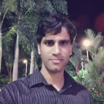 Man with dark hair wearing a striped shirt standing outdoors at night with palm trees and lights in the background.