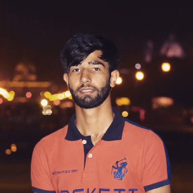Young man with dark hair and beard wearing an orange British Polo Day shirt, standing outdoors at night with blurred street lights in the background.