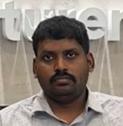 Portrait of a man with short dark hair and mustache wearing a light-colored shirt, seated in front of a blurred sign.