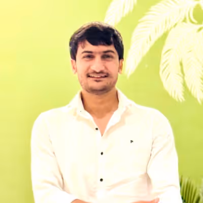 Young man with dark hair wearing a white shirt, smiling against a bright green background with leafy plants.