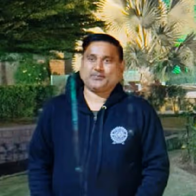 Man standing outdoors in a garden at night wearing a dark jacket with a logo on the chest.