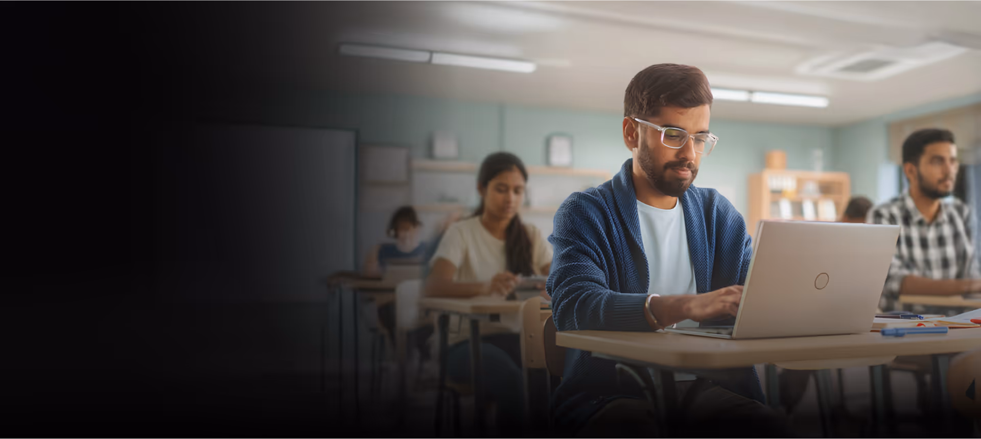 Young man wearing glasses and a blue cardigan working on a laptop in a classroom with other students in the background.