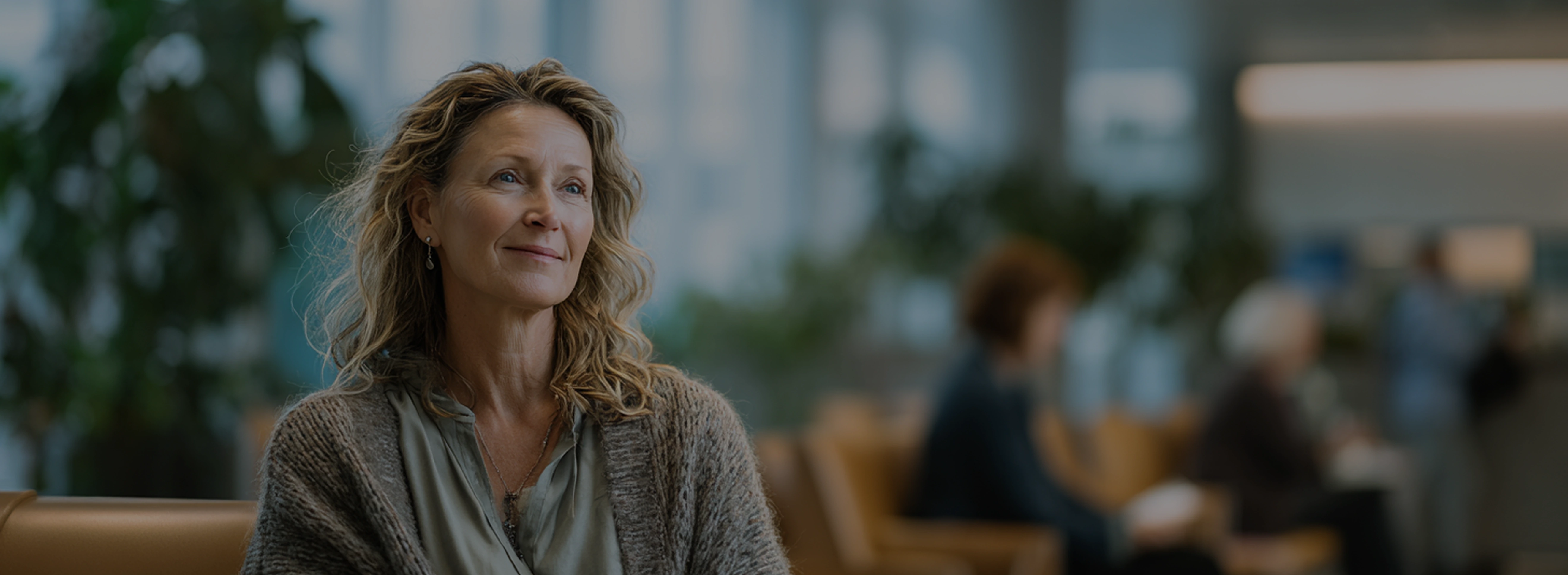 Middle-aged woman with wavy blonde hair and a knitted cardigan sitting on a bench in a softly lit waiting area.